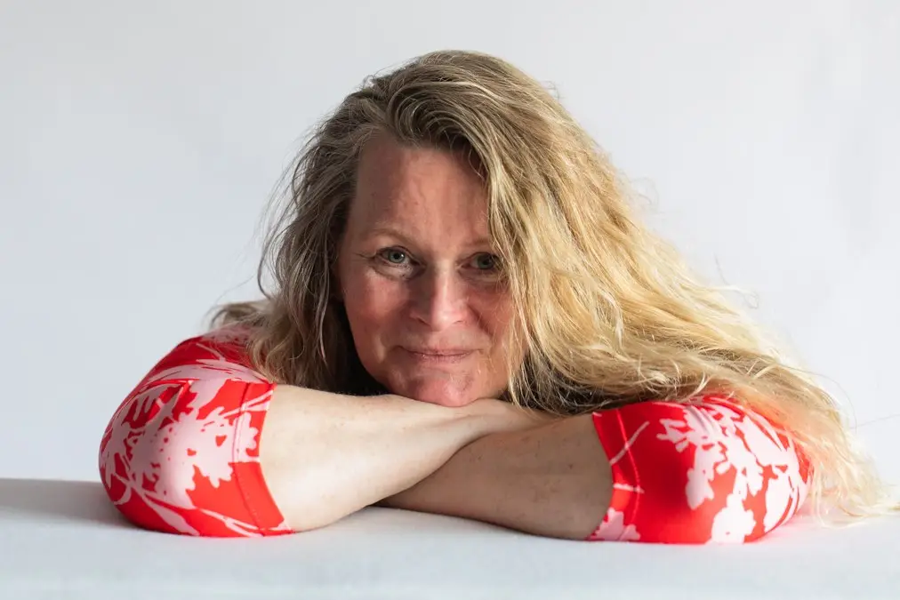 Wilma de Boer met lang blond haar in rood met witte jurk, leunend met armen op tafel en kijkend naar de camera